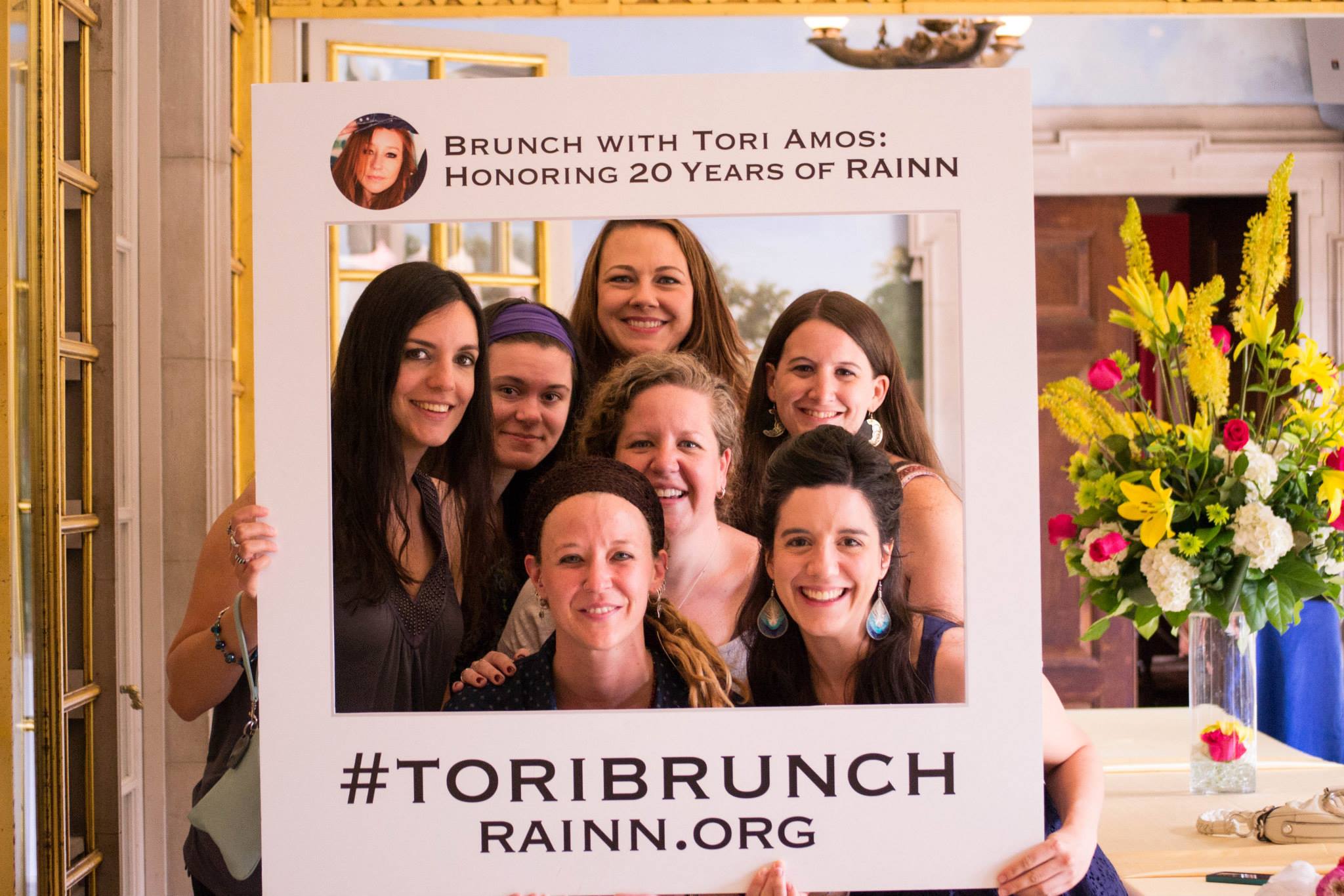 Women pose with a sign stating "Brunch with Tori Amos: Honoring 20 years of RAINN, hashtag ToriBrunch, rainn.org"